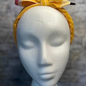 Puede incluir: Una diadema a rayas amarillas y blancas con un dise&ntilde;o de l&aacute;piz. La diadema est&aacute; atada en un lazo en la parte superior de la cabeza.
