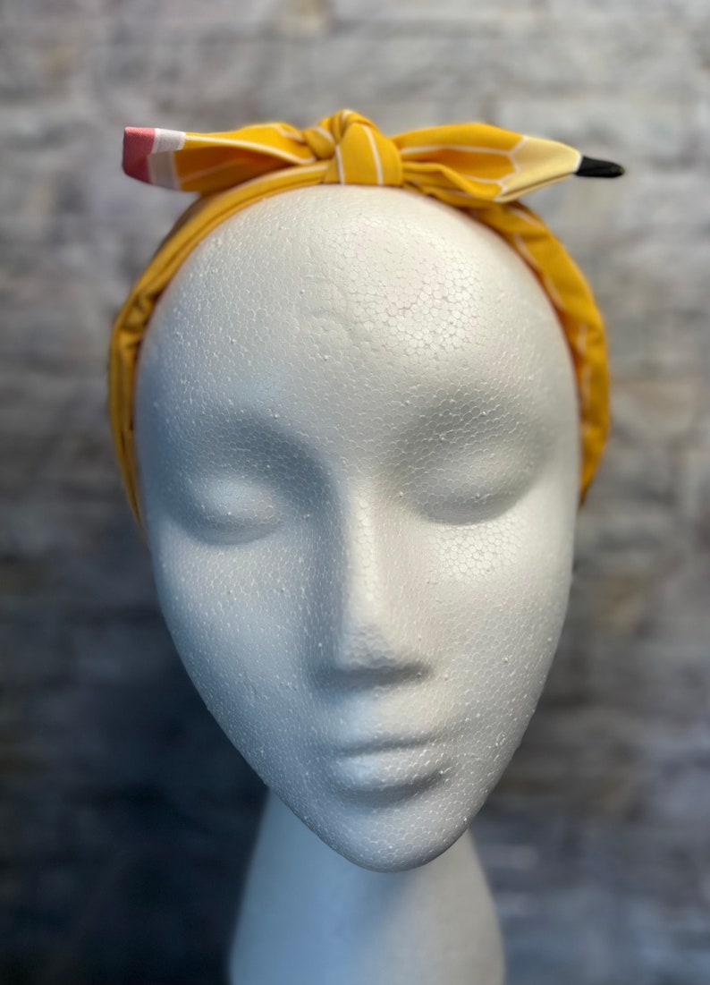 Puede incluir: Una diadema a rayas amarillas y blancas con un dise&ntilde;o de l&aacute;piz. La diadema est&aacute; atada en un lazo en la parte superior de la cabeza.