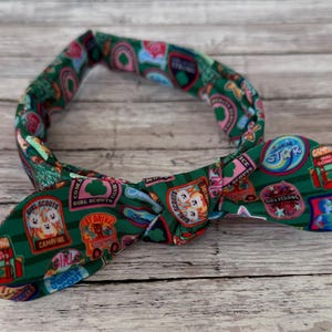 Può includere: Una fascia per capelli in tessuto verde con un motivo ripetuto di distintivi delle Girl Scout in vari colori. I distintivi includono testo come "Girl Scouts", "Cookie Star" e "Super Hero". La fascia è annodata a fiocco.