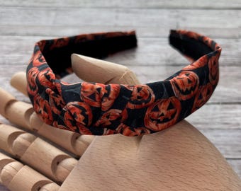 May include: A Halloween-themed headband featuring a repeating pattern of orange jack-o'-lantern faces on a black background. The headband is made of fabric and is designed to be worn on the head. The accessory is suitable for Halloween.