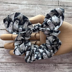 Op de afbeelding: Een zwart-witte scrunchie met een spookpatroon. De stof heeft een herhalend ontwerp van witte geesten op een donkere achtergrond. De scrunchie is geplooid en in een strikvorm geknoopt, perfect voor Halloween of thema-outfits.