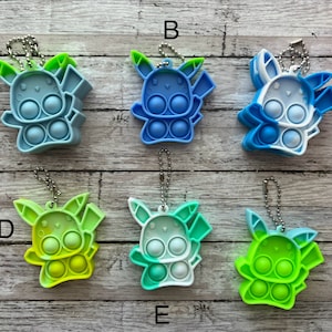 May include: Six colourful silicone Pikachu-shaped pop-it fidget toys with keychains. The toys are in various shades of blue, green, and yellow. The toys are arranged in two rows of three on a wooden surface.