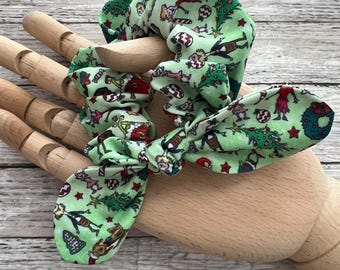 Puede incluir: Scrunchie verde con un estampado navideño con personajes de dibujos animados, bastones de caramelo y árboles de Navidad. El scrunchie está atado en un lazo.