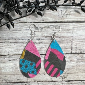 Puede incluir: Un par de pendientes en forma de lágrima con un diseño de patchwork en tonos de rosa, azul, negro y amarillo. Los pendientes están hechos de cuero y tienen ganchos plateados.