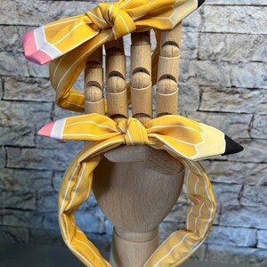 Puede incluir: Dos diademas rayadas de color amarillo y blanco con un dise&ntilde;o de l&aacute;piz. El l&aacute;piz es amarillo con una goma de borrar rosa y una punta negra.
