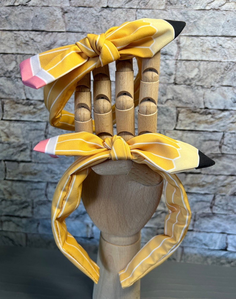 Puede incluir: Dos diademas rayadas de color amarillo y blanco con un dise&ntilde;o de l&aacute;piz. El l&aacute;piz es amarillo con una goma de borrar rosa y una punta negra.