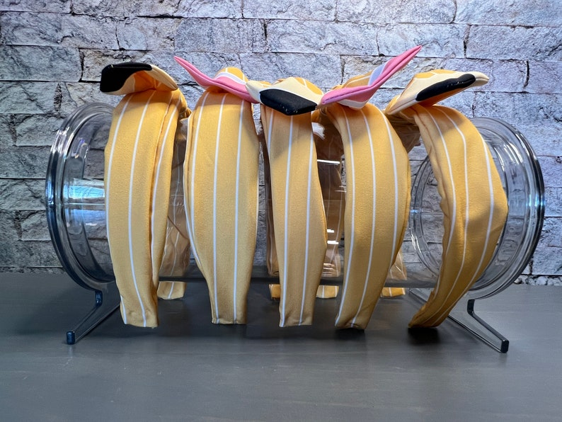 Puede incluir: Un conjunto de seis diademas de rayas amarillas y blancas con lazos de tela de diferentes colores. Las diademas se muestran en un recipiente de vidrio transparente sobre una superficie gris.