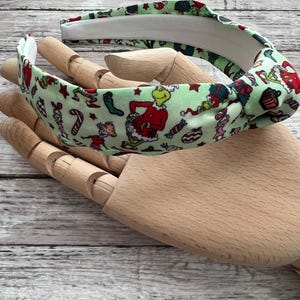 May include: Green headband with a white elastic band. The headband features a Christmas-themed print of the Grinch, candy canes, and stars.