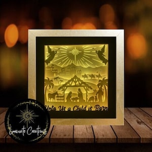 Könnte beinhalten: Ein weißer Holzrahmen mit einer beleuchteten Weihnachtskrippe im Inneren. Die Szene zeigt die Geburt Jesu mit Engeln, einem Stern und dem Text "Unto Us a Child is Born".