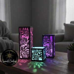 May include: Three black, geometric lanterns with tropical leaf cutouts. The lanterns are illuminated with colorful gradients of pink, purple, and green. The logo "Luminate Creations" is in the lower left corner.