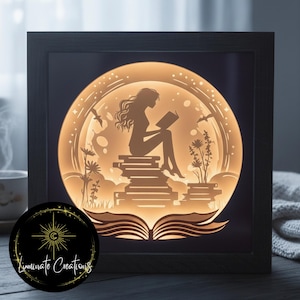 Peut inclure: Une œuvre d'art encadrée et illuminée représentant une silhouette d'une personne lisant sur une pile de livres dans un design circulaire. L'œuvre comprend des motifs floraux et d'oiseaux, avec le texte "Luminate Creations" sur un emblème circulaire.