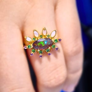 Könnte beinhalten: Ein goldener Ring mit einem großen, schillernden Opal, der von kleineren grünen und violetten Edelsteinen umgeben ist. Der Ring ist in einem Sonnenstrahl-Design mit weißen birnenförmigen Steinen gefasst.