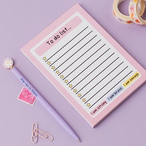 May include: Pink to-do list notepad with a white daisy pen, two rolls of washi tape, and a white ceramic pencil holder with colourful pens and scissors. The notepad has the text "To do list..." at the top and the text "I am strong I am brave I am enough" at the bottom.