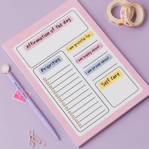 May include: A pink daily planner notepad with sections for affirmations, priorities, and self-care. Includes a purple pen, paper clips, decorative tape, and a white pen holder filled with colourful pens and pencils. The notepad has the text "Affirmation of the day".