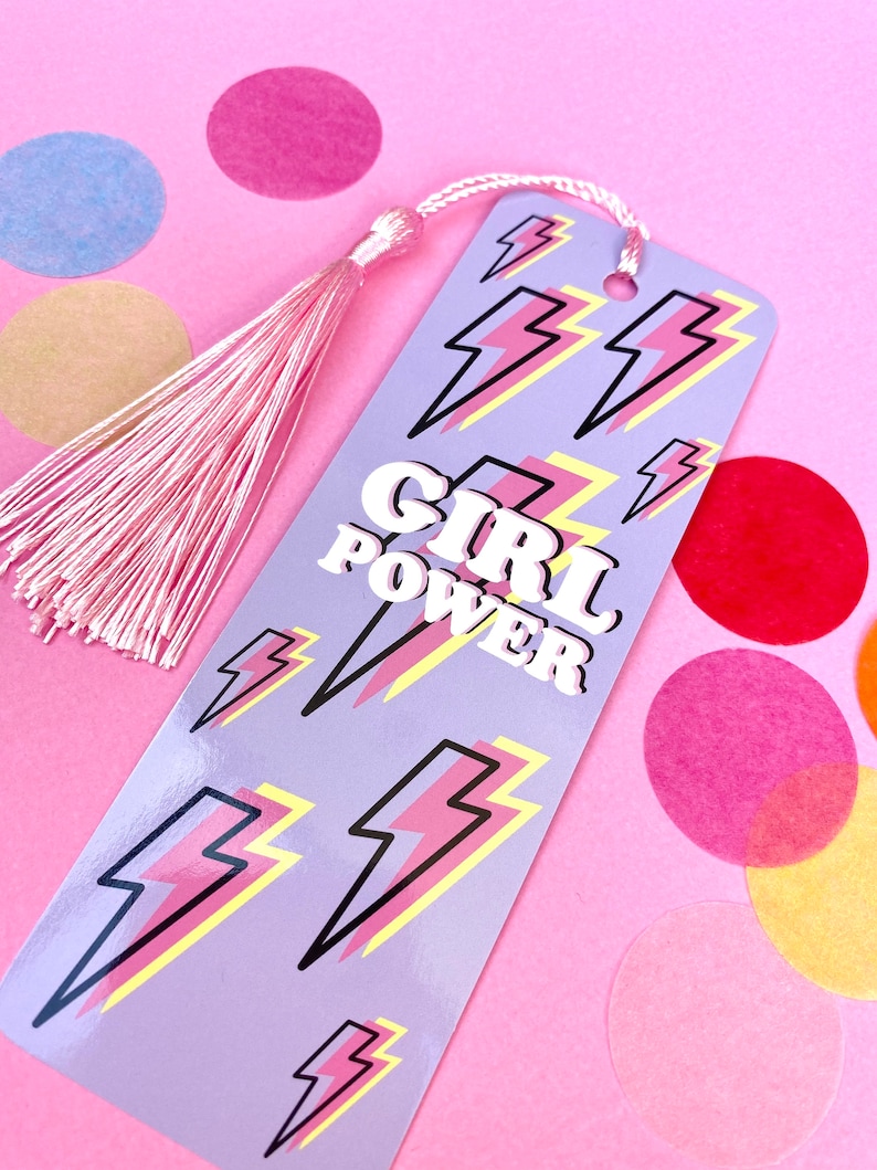 May include: A purple bookmark with a pink tassel. The bookmark has a pattern of yellow and pink lightning bolts and the words "GIRL POWER" in white letters.