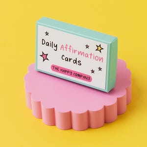 K&ouml;nnte beinhalten: Eine t&uuml;rkisfarbene Schachtel mit dem Text "Daily Affirmation Cards" und "THE HAPPY COMPANY" auf einem rosa, gewellten Sockel. Die Schachtel befindet sich auf einem gelben Hintergrund.