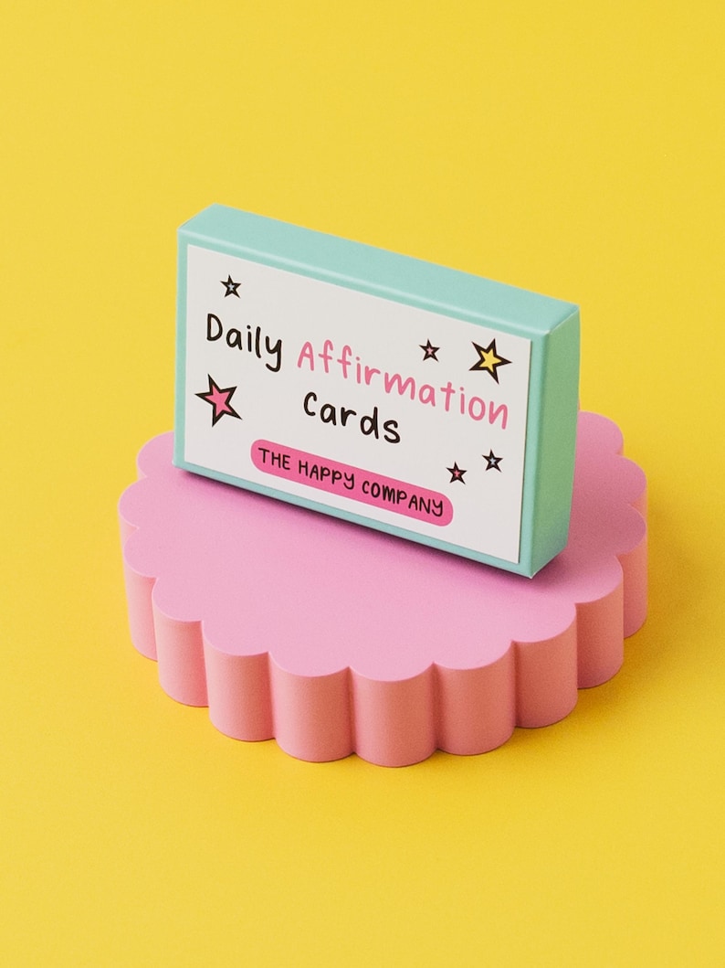 K&ouml;nnte beinhalten: Eine t&uuml;rkisfarbene Schachtel mit dem Text "Daily Affirmation Cards" und "THE HAPPY COMPANY" auf einem rosa, gewellten Sockel. Die Schachtel befindet sich auf einem gelben Hintergrund.