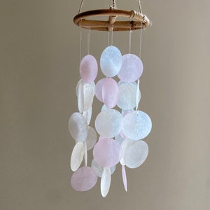 Op de afbeelding: Een windgong van witte en roze schelpen met een bamboe ring. De gong heeft meerdere lagen schelpen die aan de ring hangen.