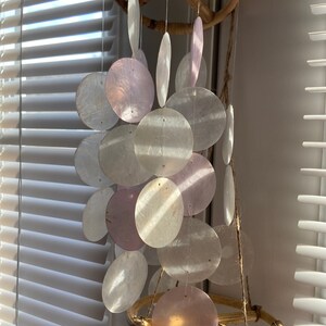 Op de afbeelding: Een windgong van witte en roze schelpen met een bamboe frame. De gong is gemaakt van veel ronde schelpen die aan een rond bamboe frame hangen.