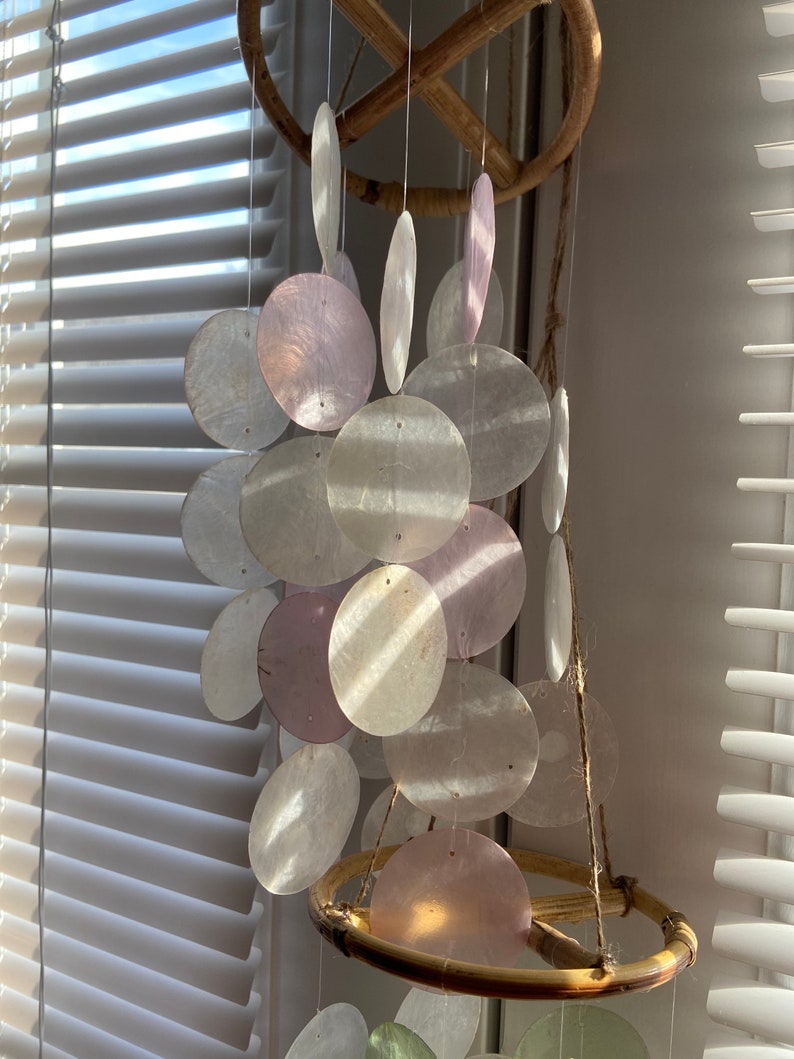 Op de afbeelding: Een windgong van witte en roze schelpen met een bamboe frame. De gong is gemaakt van veel ronde schelpen die aan een rond bamboe frame hangen.