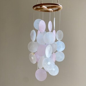 Op de afbeelding: Een windgong van witte en roze schelpen met een houten ring aan de bovenkant. De schelpen zijn in een cascade-patroon gerangschikt.