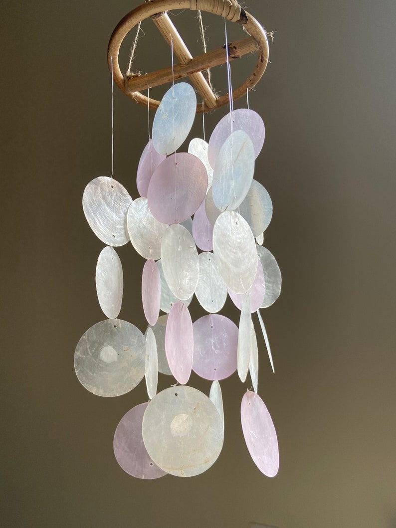 Op de afbeelding: Een windgong van witte en roze schelpen met een houten ring aan de bovenkant. De schelpen zijn aan een doorzichtig touw geregen en hangen in een watervalvorm.