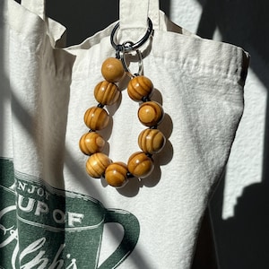 May include: A wooden bead keychain with a silver ring is attached to a white tote bag. The keychain features ten light brown, striped wooden beads. The tote bag has a green coffee cup graphic and the words "ENJOY CUP OF Khis JAVE FEE".