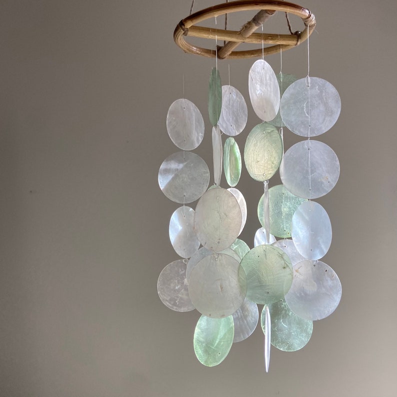 May include: A white and green wind chime made of seashells hanging from a bamboo frame. The wind chime has a variety of different sized seashells.