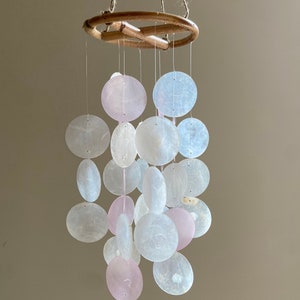 Op de afbeelding: Een windgong van witte en roze schelpen met een houten ring aan de bovenkant. De schelpen hangen aan touwtjes aan de ring.