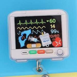 May include: A medical-themed badge reel featuring a monitor screen design. The screen displays vital signs, including a heart rate and other medical data. The badge reel is attached to a clear plastic retractable clip.