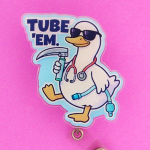 May include: A white duck wearing sunglasses, a stethoscope, and holding medical equipment, with the text "TUBE 'EM." The duck is a cartoon character on a badge reel with a clear plastic strap and a metal clip. The background is pink.