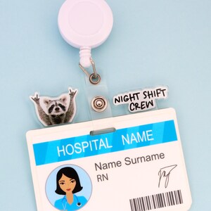 Op de afbeelding: Een witte oprolbare badge reel met een doorzichtige plastic band die een ziekenhuis-ID-badge vasthoudt. De badge heeft een blauw en wit ontwerp met de tekst "HOSPITAL NAME" en "NIGHT SHIFT CREW". Een wasbeerbedel is bevestigd.