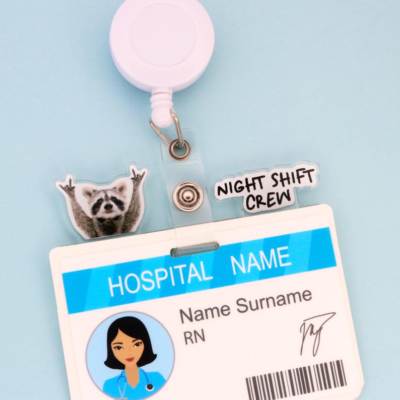 Op de afbeelding: Een witte oprolbare badge reel met een doorzichtige plastic band die een ziekenhuis-ID-badge vasthoudt. De badge heeft een blauw en wit ontwerp met de tekst "HOSPITAL NAME" en "NIGHT SHIFT CREW". Een wasbeerbedel is bevestigd.