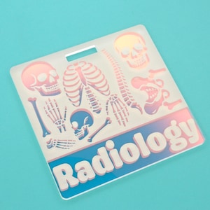 May include: A clear, rectangular badge holder with a handle, featuring a pink and blue gradient design of human skeletal elements. The word "Radiology" is printed in white block letters on a blue background.