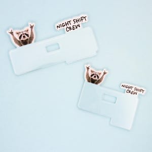 Op de afbeelding: Twee lichtblauwe telefoonhoesjes met een wasbeer die het 'rock on' handgebaar maakt. Elk hoesje heeft een wit label met de zwarte tekst 'NIGHT SHIFT CREW'. De achtergrond is effen lichtblauw.