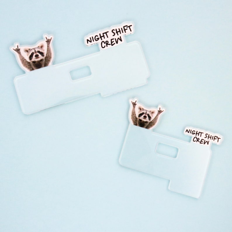 Op de afbeelding: Twee lichtblauwe telefoonhoesjes met een wasbeer die het 'rock on' handgebaar maakt. Elk hoesje heeft een wit label met de zwarte tekst 'NIGHT SHIFT CREW'. De achtergrond is effen lichtblauw.