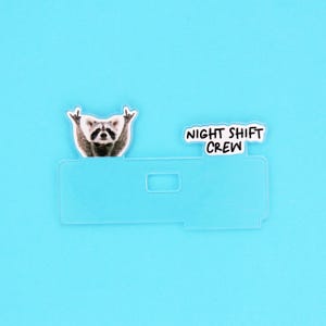 Op de afbeelding: Een doorzichtige plastic kaarthouder met een wasbeer-afbeelding en de tekst "NIGHT SHIFT CREW" op een blauwe achtergrond. De wasbeer maakt een rock-and-roll handgebaar.