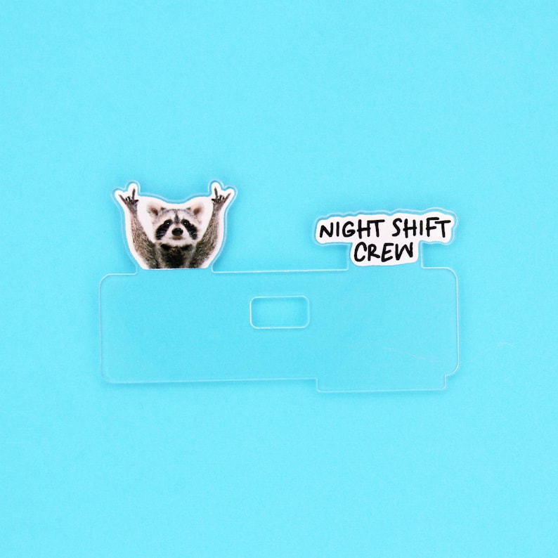 Op de afbeelding: Een doorzichtige plastic kaarthouder met een wasbeer-afbeelding en de tekst "NIGHT SHIFT CREW" op een blauwe achtergrond. De wasbeer maakt een rock-and-roll handgebaar.