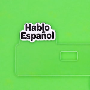 May include: A clear acrylic sign featuring a white die-cut sticker with the black text Hablo Español in a bold, rounded sans-serif font. The sign has a rectangular cutout on the right side and a smooth, transparent finish throughout the piece.