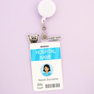 Op de afbeelding: Een witte intrekbare badge reel met een doorzichtige plastic ID-houder. De ID heeft een blauwe streep met "HOSPITAL NAME" erop gedrukt. Een wasbeerbedel en de woorden "NIGHT SHIFT CREW" zijn ook bevestigd.