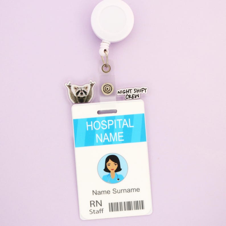 Op de afbeelding: Een witte intrekbare badge reel met een doorzichtige plastic ID-houder. De ID heeft een blauwe streep met "HOSPITAL NAME" erop gedrukt. Een wasbeerbedel en de woorden "NIGHT SHIFT CREW" zijn ook bevestigd.