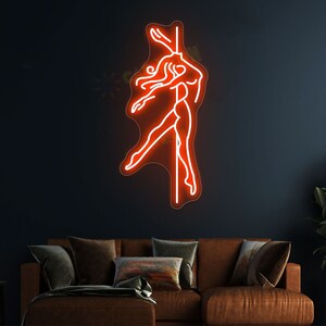 K&ouml;nnte beinhalten: Ein Neon-Schild, das eine Frau in Silhouette zeigt, die an einer Stange tanzt. Das Schild leuchtet in einem leuchtenden Orange und ist an einer dunklen Wand montiert.