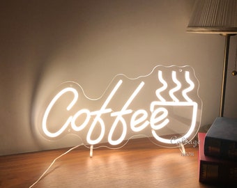 May include: White neon sign with the word "Coffee" and a stylized image of a steaming coffee cup. The sign is on a wooden surface with a lamp and books in the background.
