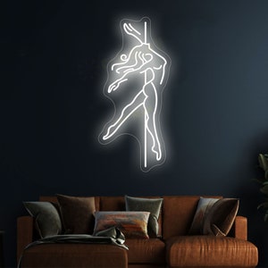 K&ouml;nnte beinhalten: Ein wei&szlig;es Neon-Schild, das eine Frau in Silhouette zeigt, die auf einer Stange posiert. Das Schild ist an einer dunkelblauen Wand &uuml;ber einem braunen Sofa angebracht.