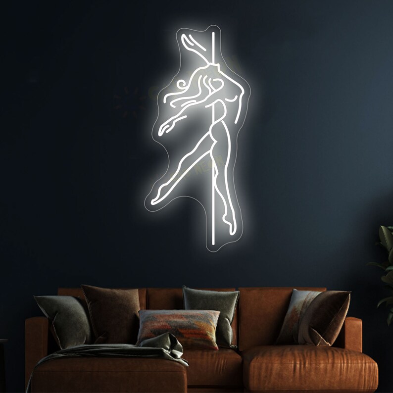 K&ouml;nnte beinhalten: Ein wei&szlig;es Neon-Schild, das eine Frau in Silhouette zeigt, die auf einer Stange posiert. Das Schild ist an einer dunkelblauen Wand &uuml;ber einem braunen Sofa angebracht.