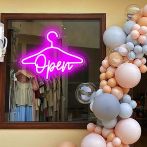 May include: Pink neon sign shaped like a clothes hanger with the word "Open" in a cursive font. The sign is hanging in a storefront window with clothing displayed behind it. A garland of pastel balloons is in the foreground.