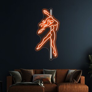 K&ouml;nnte beinhalten: Ein orangefarbenes Neon-Schild, das eine Frau zeigt, die an einer Stange tanzt. Das Schild ist an einer dunklen Wand &uuml;ber einem braunen Ledersofa angebracht.