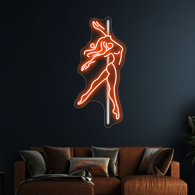 K&ouml;nnte beinhalten: Ein orangefarbenes Neon-Schild, das eine Frau zeigt, die an einer Stange tanzt. Das Schild ist an einer dunklen Wand &uuml;ber einem braunen Ledersofa angebracht.