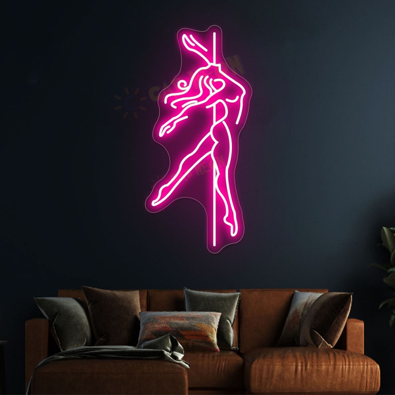 K&ouml;nnte beinhalten: Pinkfarbenes Neon-Schild, das eine Frau in einer T&auml;nzerpose an einer Stange zeigt. Das Schild ist an einer dunklen Wand &uuml;ber einem braunen Sofa angebracht.