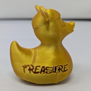 以下が含まれることがあります： 前面に「TREASURE」の文字が刻まれた金色のアヒルの置物。アヒルは小さな帽子をかぶっており、光沢のある金属素材でできています。この置物は装飾品です。